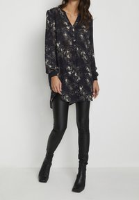 Blusa nera a maniche lunghe con un motivo floreale in colori tenui, dotata di una fila di bottoni e un orlo asimmetrico, abbinata a leggings neri lucidi.