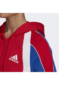 Sudadera roja con cremallera y acentos en franjas blancas y azules. Presenta una capucha, una textura suave y un logo de Adidas en blanco en la parte frontal.