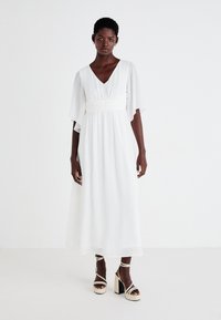 Λευκό, αέρινο φόρεμα με V-λαιμόκοψη, φαρδιές μανσέτες και σφιχτή μέση. Συνδυασμένο με μπεζ πλατφόρμες με λωρίδες που δένουν στον αστράγαλο.