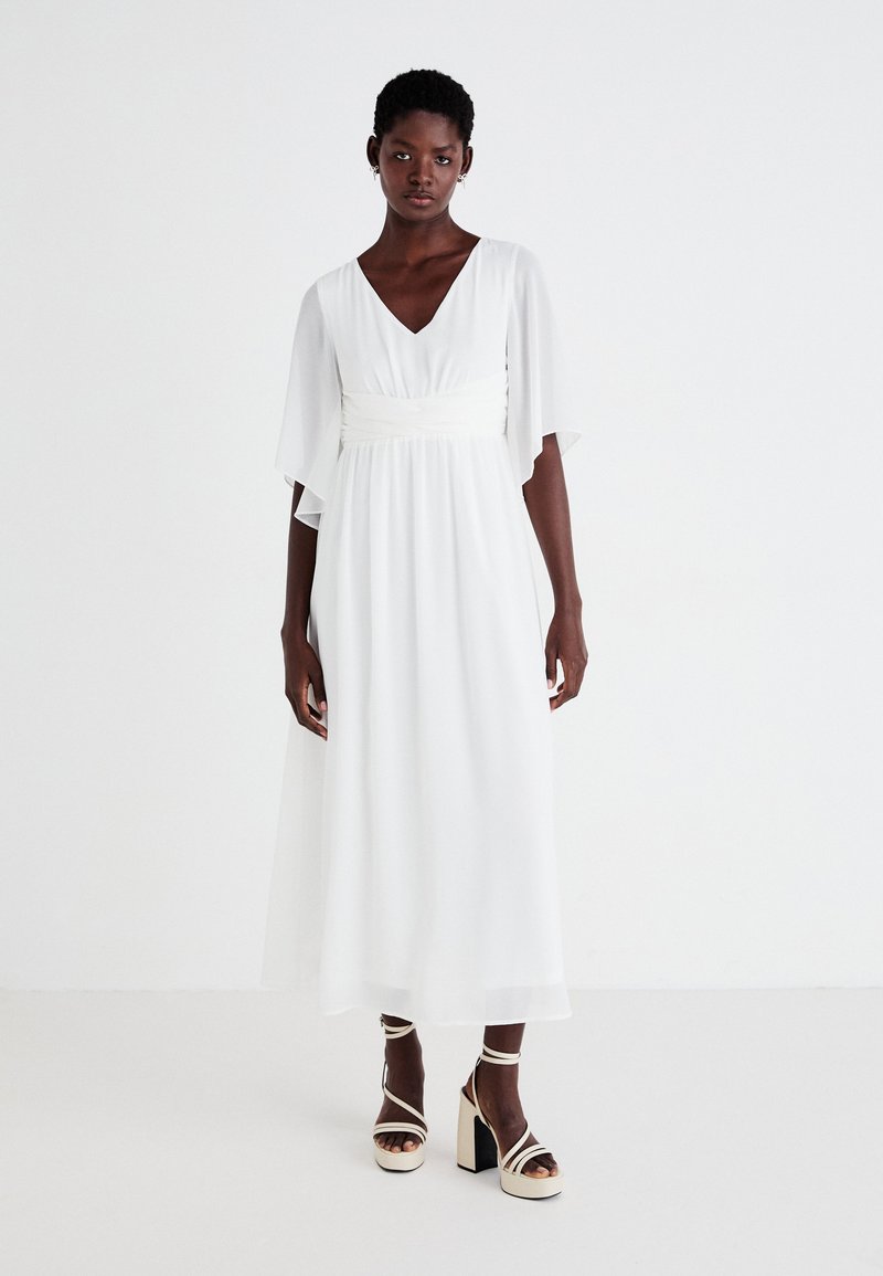 Λευκό, αέρινο φόρεμα με V-λαιμόκοψη, φαρδιές μανσέτες και σφιχτή μέση. Συνδυασμένο με μπεζ πλατφόρμες με λωρίδες που δένουν στον αστράγαλο.