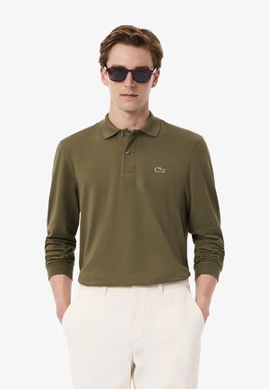 Camisa polo de manga larga verde oliva con un collar clásico, tres botones y un pequeño logo de cocodrilo bordado en el lado izquierdo del pecho.