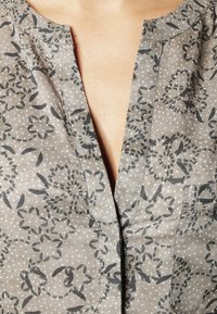Grå stofbluse med blomsterprint og polkaprikker, med en V-hals og knaplukning foran. Blødt tekstur med synlige sømme.