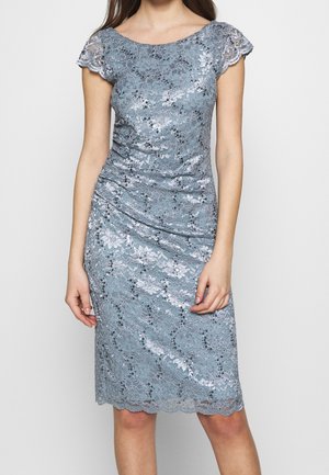 Mujer con un vestido ajustado de encaje azul plateado, con mangas cortas con borde ondulado y detalles de lentejuelas, posando frente a un fondo claro y sencillo.