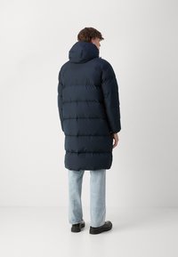 Man wearing a long black padded coat with hood, light blue jeans, and black shoes, standing facing away against a plain background.