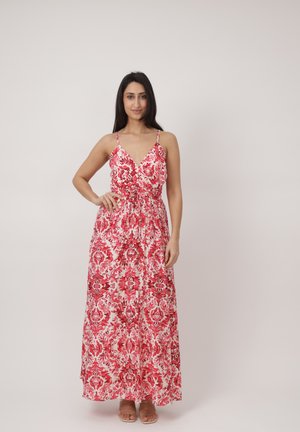 Kobieta stojąca w czerwono-białej sukience maxi w kwiaty bez rękawów z cienkimi ramiączkami na jednolitym jasnym tle.