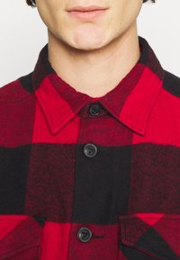 Camicia a quadri rossa e nera con bottone, caratterizzata da un colletto, bottoni neri e un tessuto morbido e texturizzato con due tasche sul petto.