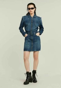 Denimmekko pitkillä hihoilla, vetskari edessä ja vyötäröllä vyö. Tummansininen väri, kevyt kulutushelma. Yhdistetty mustiin nilkkap bootseihin.