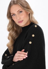 Maglione nero lavorato a maglia con collo alto, dotato di bottoni dorati lungo la spalla. Texture morbida e vestibilità rilassata messe in risalto.