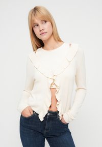 Ξανθιά γυναίκα που φοράει ένα κρεμ ζακέτα με βολάν πάνω από ένα ταιριαστό τοπ και σκούρα μπλε τζιν, με τα χέρια στις τσέπες, κοιτάζει προς το πλάι.