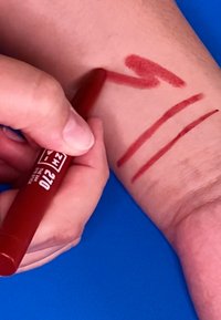 Red eyeliner pencil applies swatches on a light skin tone arm with curved and straight lines; backdrop is a solid blue surface.