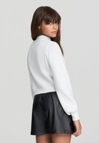 Maglione bianco corto con collo alto, maniche lunghe a palloncino e orlo a coste. Abbinato a pantaloni shorts in pelle nera. Texture liscia, design minimale.