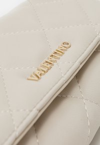 Cartera de cuero acolchado beige con hardware de logo dorado, costuras prominentes y una superficie texturizada con un diseño limpio.