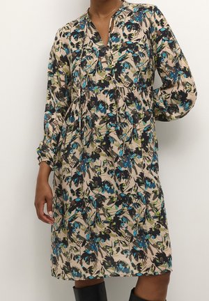 Žena v béžových šatech po kolena s modrým, černým a zeleným květinovým potiskem, dlouhými rukávy a výstřihem do V, v kombinaci s černými kozačkami.