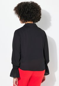 Schwarze Bluse mit langen, gepufften Ärmeln und elastischen Bündchen. Der Stoff ist leicht und durchscheinend und wird mit roten Hosen kombiniert.