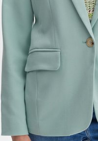 Mint green blazer with a textured finish, single button closure, and a decorative pocket on the left side. Tailored fit.