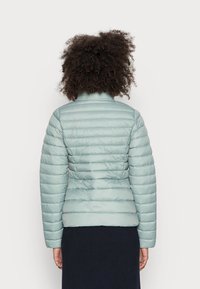 Doudoune bleu clair avec un design matelassé horizontal, col montant et manches longues. Tissu texturé, silhouette ajustée.