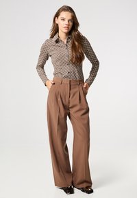Bruine gestreepte shirt met een puntkraag, gecombineerd met hoge, wijde bruine broek. Model draagt donkere schoenen. Neutrale achtergrond.
