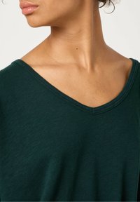 Tumši zaļa v-veida krekls, izgatavots no mīkstas, vieglas auduma; tam ir brīva piegriezuma forma un nemanāma dūriena ap kakla izgriezumu.