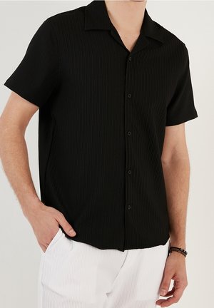 Man draagt een zwart kortere mouwen overhemd met textuur en witte textuurbroeken, met hand in de zak, tegen een effen achtergrond.