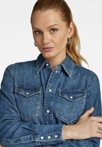 Camicia in denim di colore blu medio con bottoni frontali, due tasche sul petto e dettagli in metallo argentato. Presenta un colletto classico e polsini a scatto.