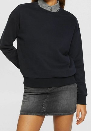 Personne portant un sweat-shirt noir par-dessus une chemise grise à col, associée à une mini-jupe en denim gris foncé, debout avec une main derrière le dos.