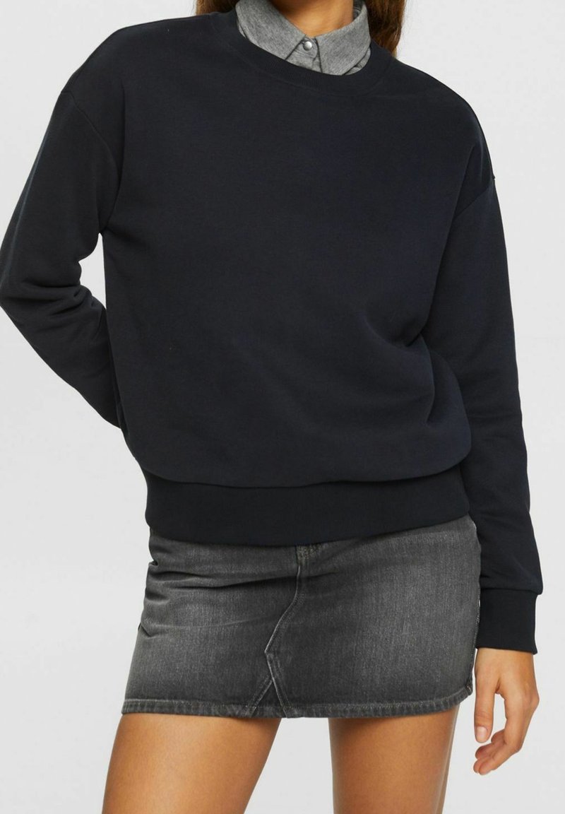 Personne portant un sweat-shirt noir par-dessus une chemise grise à col, associée à une mini-jupe en denim gris foncé, debout avec une main derrière le dos.