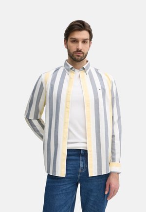 Camisa de algodón a rayas con rayas verticales en gris, blanco y amarillo, con cuello abotonado y mangas largas. Usada sobre una camiseta blanca.
