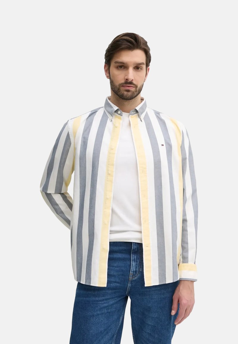 Camisa de algodón a rayas con rayas verticales en gris, blanco y amarillo, con cuello abotonado y mangas largas. Usada sobre una camiseta blanca.