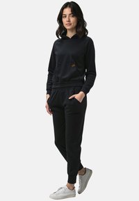 Černá mikina s kapucí, přední kapsou a pružnými manžetami, doplněná o černé tepláky. Bílé tenisky dotvářejí celý outfit.