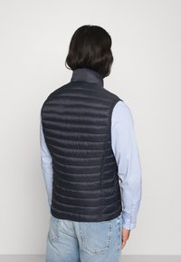 Gilet puffer navy con colletto alto, caratterizzato da una texture trapuntata, materiale leggero e una vestibilità aderente, indossato sopra una camicia a quadri blu.