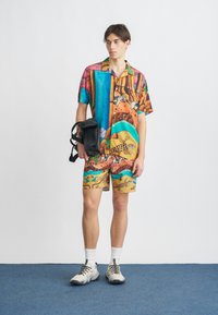 Completo con camicia a maniche corte e pantaloncini in motivi multicolori vivaci. Realizzato con tessuto leggero con motivi tropicali. Indossato con una borsa nera.