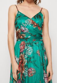 Floral dress in teal fabric features a V-neckline, thin straps, and a fitted bodice with a flowing skirt adorned with multicolored flowers.