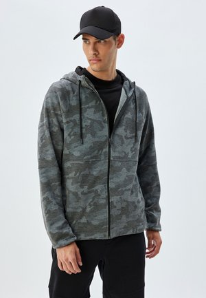 Grå camouflage hættetrøje med lynlås foran, to sidelommer og snørresnor til hætten, båret over en sort t-shirt og kombineret med sorte bukser.