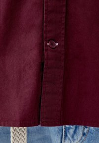 Diep bordeauxrood shirt met een doorgeknoopte placket en contrasterende stiksels, met een zachte textuur en een zoom net boven een blauwe denimachtergrond.