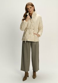 Frau in einer beigen gesteppte Jacke mit Kordelzügen, karierten weiten Hosen und braunen Knöchelstiefeln, posiert vor einem einfarbigen hellen Hintergrund.