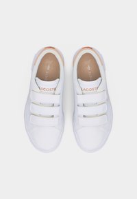 Λευκά αθλητικά παπούτσια Lacoste με τρεις λουριά βέλκρο και καφέ εσωτερικές σόλες, απεικονισμένα από πάνω σε απλό φόντο.