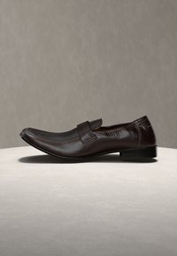 Καφέ σκούρο δερμάτινο μοκασίνι ανδρικό με χαμηλό τακούνι και λεπτομέρεια με λουράκι στο πάνω μέρος, εμφανίζεται σε ουδέτερη επιφάνεια.