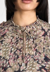 Bluză cu imprimeu floral dintr-un material moale, având un decolteu ondulat și un șnur legat cu accente metalice. Culorile includ bej, roz și negru.