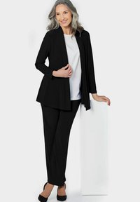 Schwarze Langarmjacke über einem weißen Oberteil mit Verzierungen, kombiniert mit schwarzen Hosen und schwarzen High Heels. Einfaches, maßgeschneidertes Design.