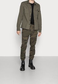 Uomo che indossa una giacca verde oliva con bottoni, una maglietta nera, pantaloni mimetici con cordoncino e stivali neri in pelle alla caviglia su un pavimento grigio.