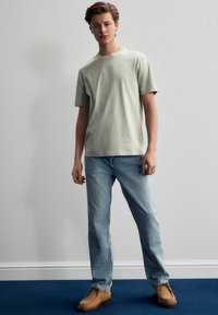 Hellgrünes Baumwoll-T-Shirt mit Rundhalsausschnitt und kurzen Ärmeln, kombiniert mit hellblauen Jeans und braunen, strukturierten Schuhen in einer minimalistischen Umgebung.