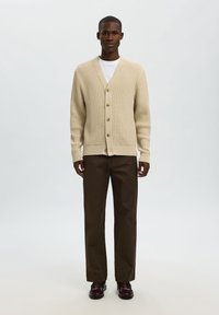 Beżowy sweter zapinany na guziki w serek z pięcioma guzikami, zestawiony z brązowymi spodniami. Tekstura jest prążkowana, a krój luźny. Brązowe buty eleganckie.