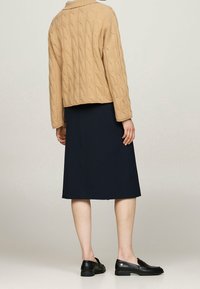 Pull en tricot beige avec un col, assorti à une jupe bleu marine mi-longue et des mocassins noirs. Accent sur la texture et le design superposé.