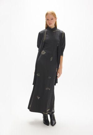 Mujer con pelo rojo liso que lleva un vestido negro largo con tachuelas doradas y pequeños estampados gráficos, combinado con botines negros.