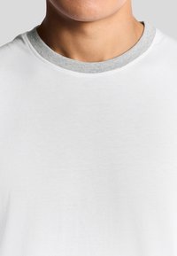 T-shirt in cotone bianco con scollo a coste grigio. Il tessuto appare morbido e liscio, mostrando una leggera texture. Nessuna grafica o motivo.