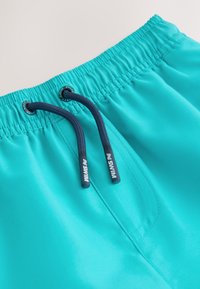 Türkiissinised ujumispüksid elastse vöökohaga, tumesinised nöörid ning brändi detailid otsadel. Sile, kerge kangas korraliku õmblusega.