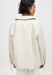 Kreminė odinė striukė laisvu kirpimu. Yra plati apykaklė, ilgomis rankovėmis su užtrauktukais ir lygi tekstūra. Rodyti atgalinį vaizdą.