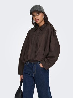 Vrouw draagt een losse bruine jas, donkerblauwe spijkerbroek, grijze pet en houdt een zwarte handtas vast tegen een effen lichte achtergrond.