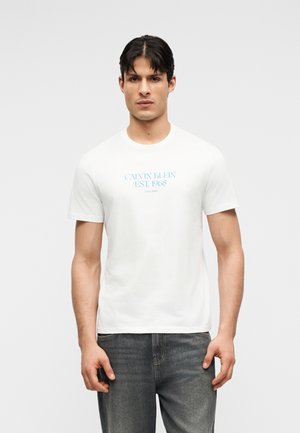 Νεαρός άνδρας με κοντά σκούρα μαλλιά, φοράει λευκό μπλουζάκι Calvin Klein με μπλε γράμματα και γκρι τζιν, στέκεται μπροστά σε απλό φόντο.