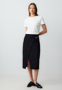 Zwarte wikkelrok met een hoge split, voorzien van een gouden accent. Gecombineerd met een witte aansluitende t-shirt en zwarte schoenen. Soepele textuur.
